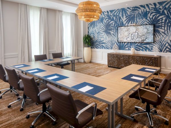 A conference room with U-shaped tables, brown chairs, blue accents, and a decorative wall, under a wicker chandelier.