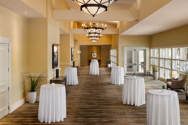 This image shows a hallway with tables covered in white cloths, chairs by windows, and chandeliers above, likely set for an event.
