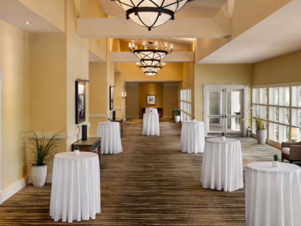 This image shows a hallway with tables covered in white cloths, chairs by windows, and chandeliers above, likely set for an event.
