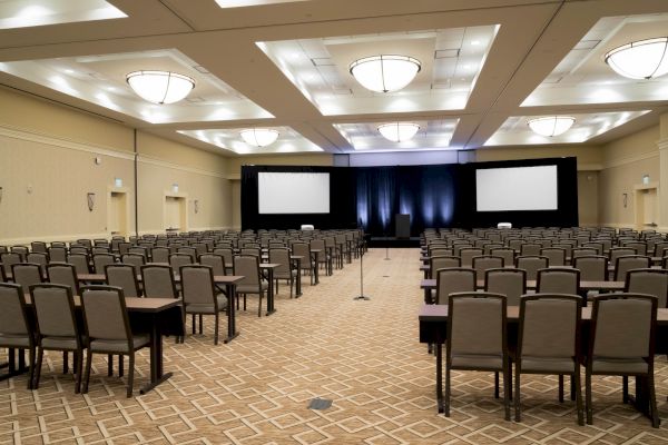 An empty conference room with rows of chairs and tables facing a stage with two large screens and lights.