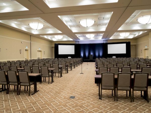 An empty conference room with rows of chairs and tables facing a stage with two large screens and lights.