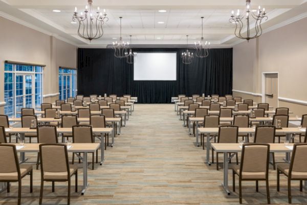 An empty conference room with rows of tables and chairs facing a projector screen, featuring chandeliers and neutral decor.
