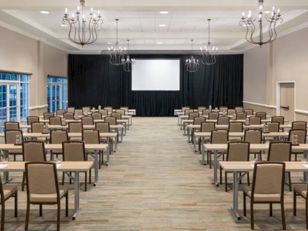 An empty conference room with rows of tables and chairs facing a projector screen, featuring chandeliers and neutral decor.