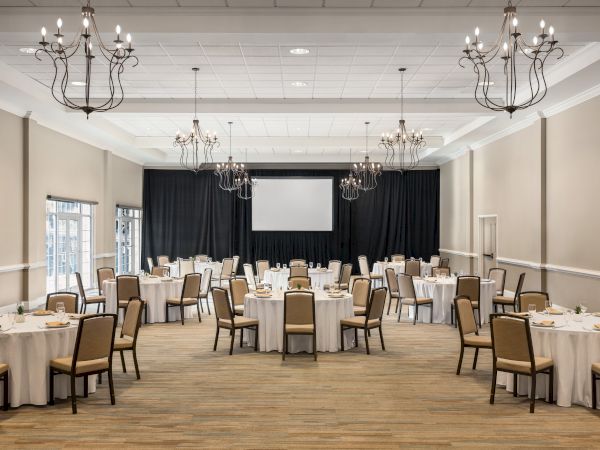 A banquet hall with round tables set for a formal event, chandeliers on the ceiling, and a large projector screen at the front.