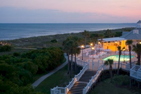 A beachside venue at dusk with palm trees, a lit pool, and a pathway leading to the ocean, creating a tranquil and inviting scene.