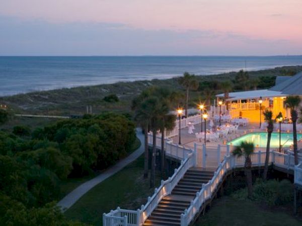 A beachside venue at dusk with palm trees, a lit pool, and a pathway leading to the ocean, creating a tranquil and inviting scene.