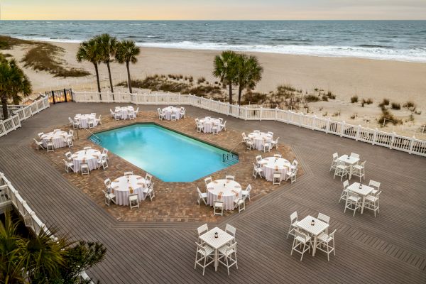 A beachfront setting with a pool, surrounded by tables and chairs on a wooden deck, with palm trees and ocean in the background.