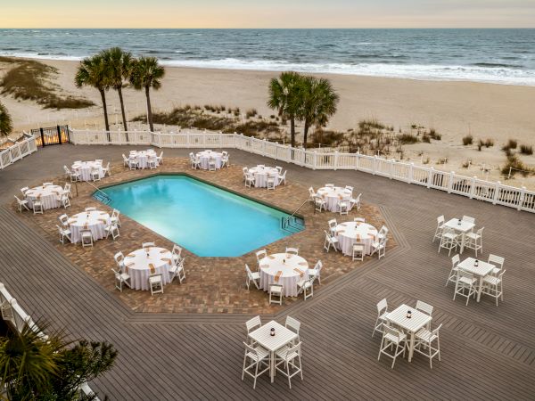 A beachfront setting with a pool, surrounded by tables and chairs on a wooden deck, with palm trees and ocean in the background.