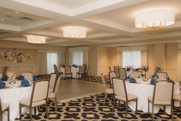 The image shows an elegant banquet hall with round tables, chairs, and blue napkins arranged for an event.