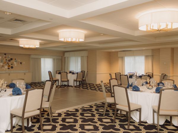 The image shows an elegant banquet hall with round tables, chairs, and blue napkins arranged for an event.