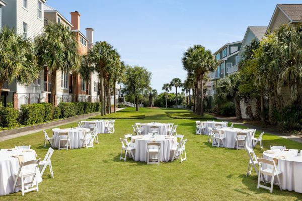 Outdoor setup with round tables and white chairs on a grassy area, surrounded by palm trees and buildings.