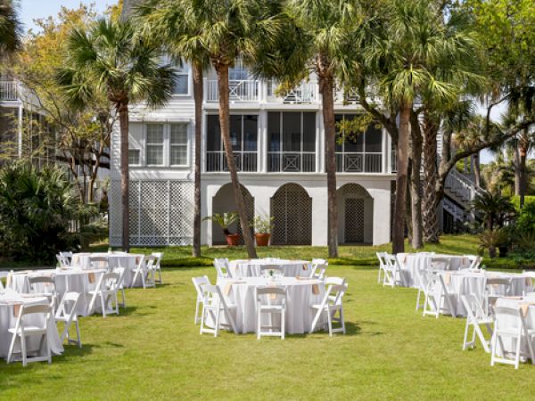 Outdoor setting with round tables and white chairs on a lawn, surrounded by palm trees and houses in the background.