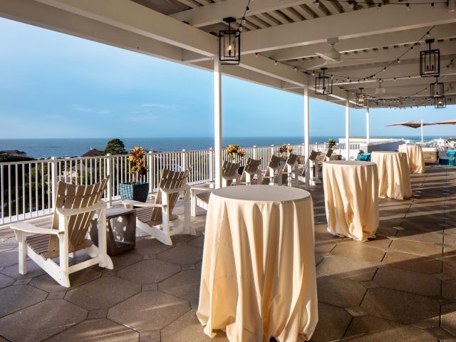 An outdoor terrace with white chairs, round tables with cloths, potted plants, and ocean views under a roof with string lights and lanterns.