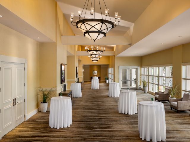 A well-lit hallway with chandeliers, round tables with white cloths, potted plants, and large windows providing natural light.
