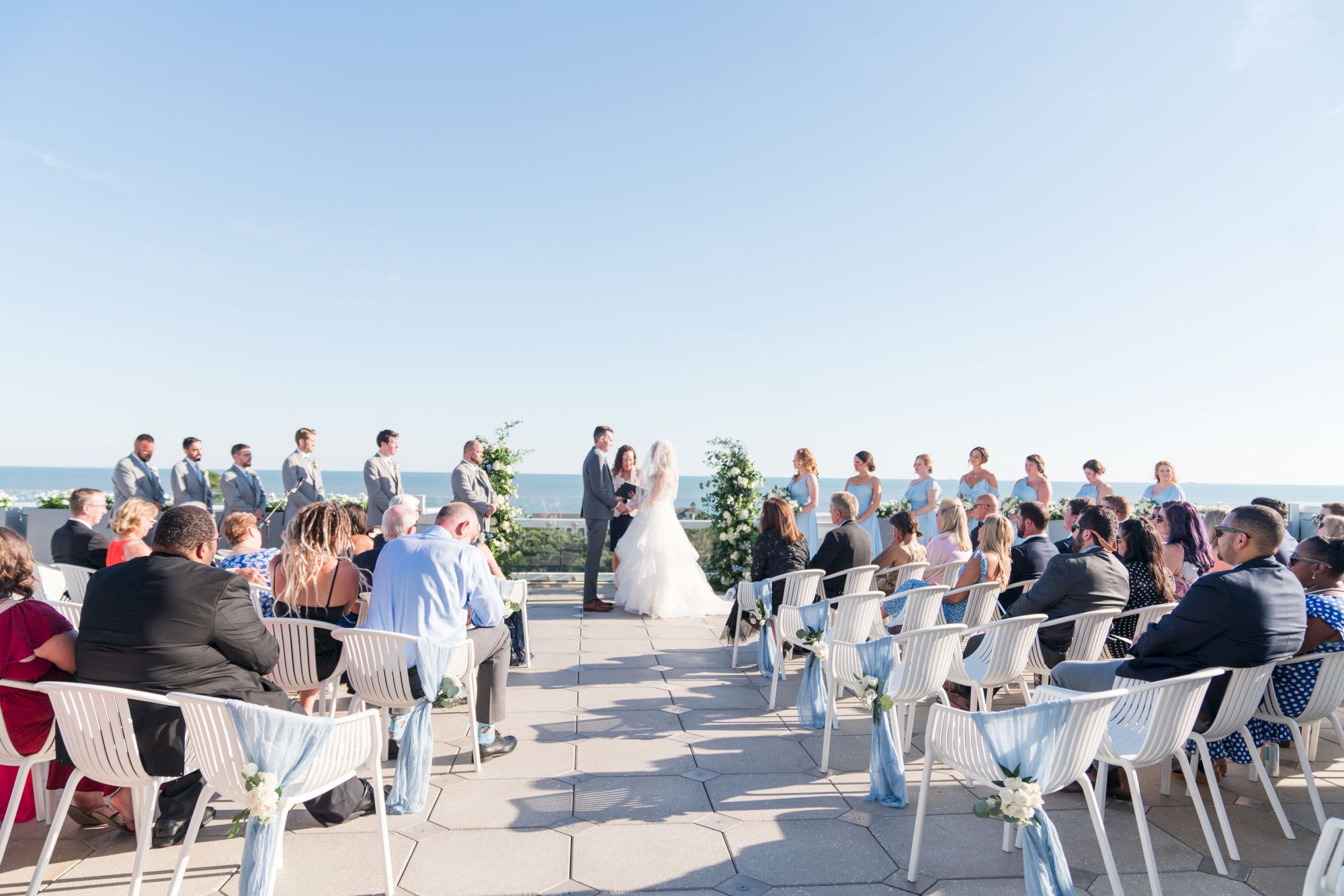 A wedding ceremony outdoors with a couple at the altar, bridesmaids, groomsmen, and seated guests on a sunny day.