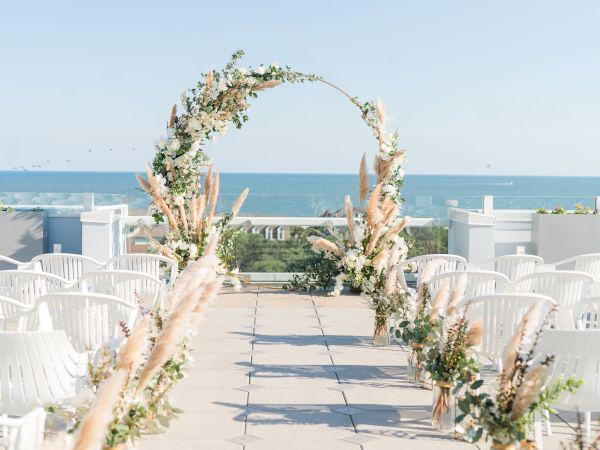 An outdoor wedding setup with a floral archway, white chairs, and ocean view.