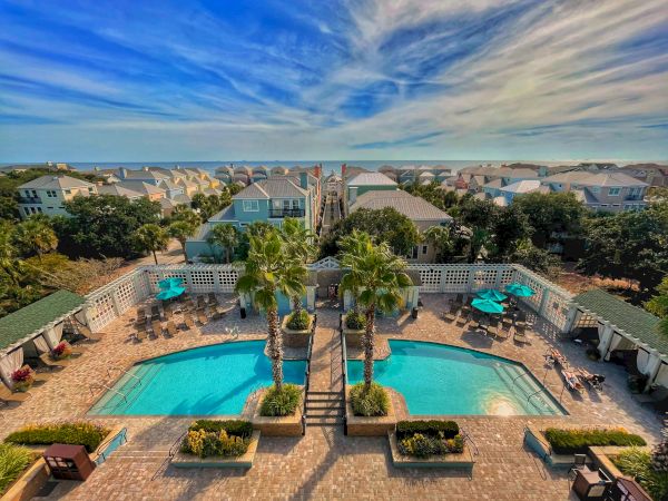Aerial view of a resort with two pools surrounded by palm trees, lounge chairs, and cabanas, with houses and blue sky in the background.