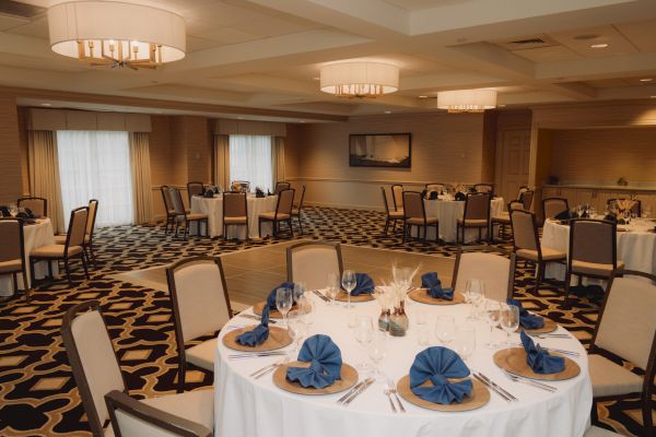 An elegantly set banquet room with round tables, blue napkins, wine glasses, and overhead lighting, creating a formal dining atmosphere.