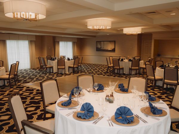 An elegantly set banquet room with round tables, blue napkins, wine glasses, and overhead lighting, creating a formal dining atmosphere.