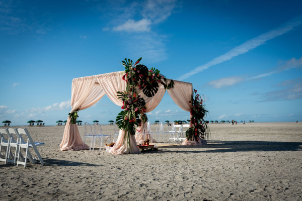 A beach setup featuring a decorated canopy with drapes and arranged chairs, possibly for a wedding or event, under a clear sky.