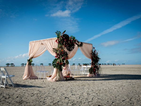 A beach setup featuring a decorated canopy with drapes and arranged chairs, possibly for a wedding or event, under a clear sky.