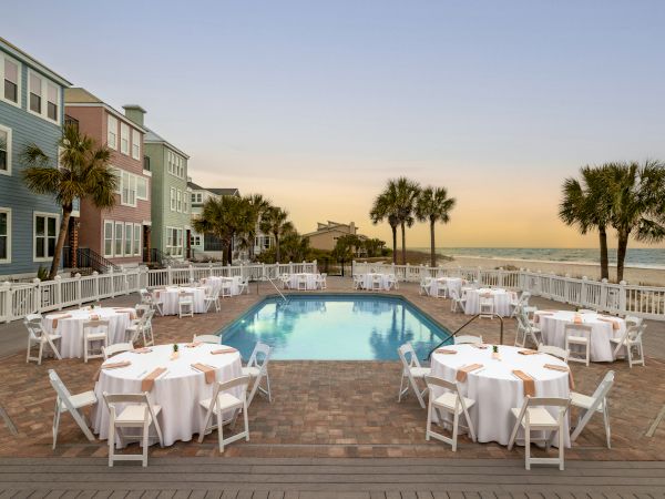 A beachside setup with round tables covered in white cloth surrounding a pool, palm trees, colorful houses, and ocean view at sunset.