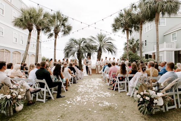 A beachside wedding ceremony with guests seated on either side; a couple stands at the altar under palm trees and string lights.