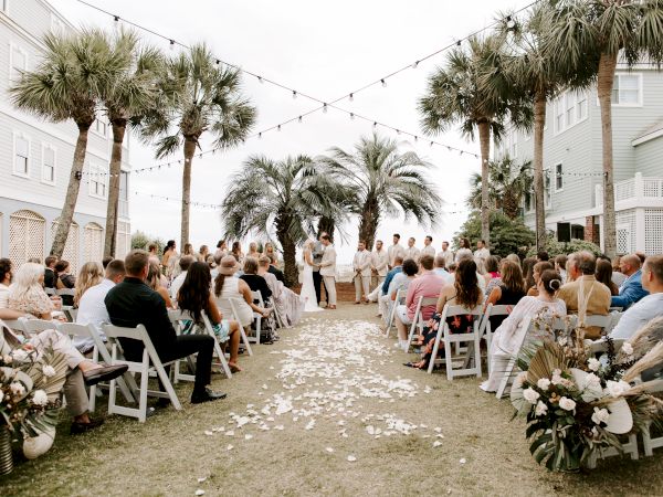 A beachside wedding ceremony with guests seated on either side; a couple stands at the altar under palm trees and string lights.