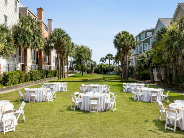 Outdoor event setup with round tables and white chairs on grass, surrounded by palm trees and buildings under a clear blue sky.