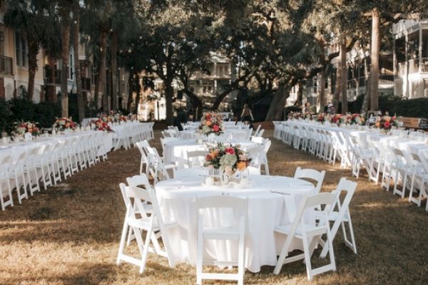 An outdoor event setup with white tables and chairs, adorned with floral centerpieces, arranged in a garden setting.