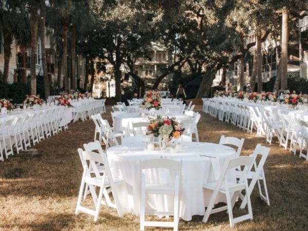 An outdoor event setup with white tables and chairs, adorned with floral centerpieces, arranged in a garden setting.