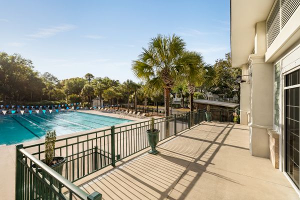 A swimming pool with lane markers, surrounded by palm trees and lounge chairs, viewed from a balcony with railing and potted plants.