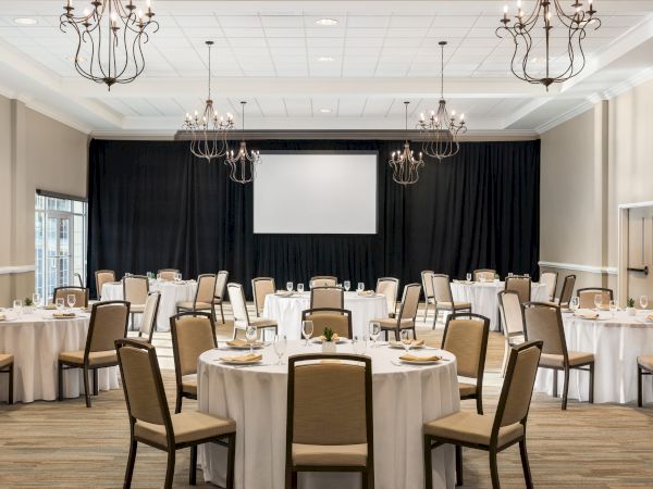 A banquet hall with round tables set for an event, featuring chandeliers, a projector screen, and neutral decor.