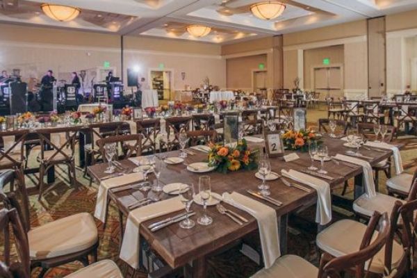 The image shows a banquet hall set for an event with tables, chairs, place settings, floral arrangements, and a stage with a band setup.