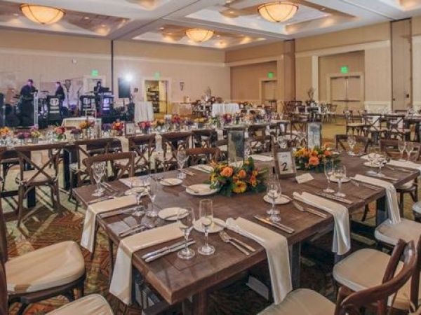 The image shows a banquet hall set for an event with tables, chairs, place settings, floral arrangements, and a stage with a band setup.