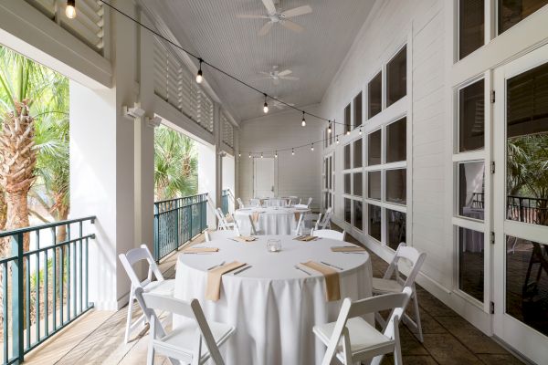 A covered patio with round tables set for an event, featuring white tablecloths, string lights, and outdoor views through windows.