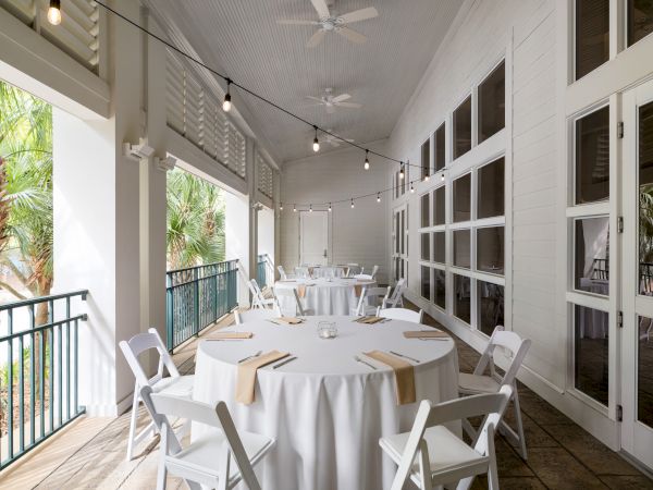 A covered patio with round tables set for an event, featuring white tablecloths, string lights, and outdoor views through windows.
