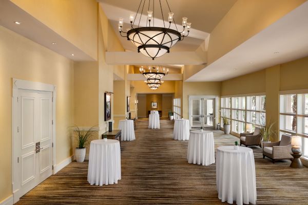 A spacious hallway with elegant chandeliers, high tables covered in white cloth, large windows, and cozy seating.