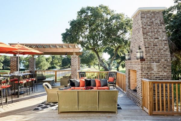 The image shows an outdoor patio with seating, a red brick fireplace, umbrella-covered tables, and trees in the background.