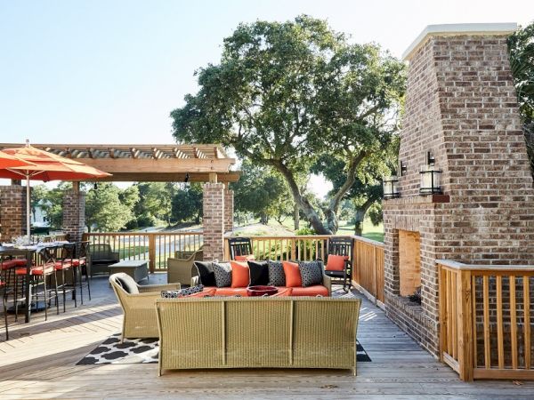 The image shows an outdoor patio with seating, a red brick fireplace, umbrella-covered tables, and trees in the background.