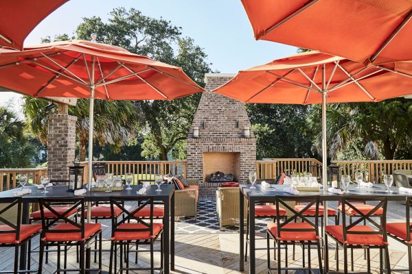 Outdoor dining area with orange umbrellas, tables set with glasses, surrounded by trees, and a brick fireplace in the background.