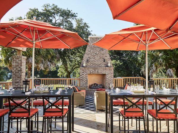 Outdoor dining area with orange umbrellas, tables set with glasses, surrounded by trees, and a brick fireplace in the background.