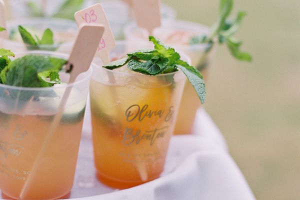 The image shows plastic cups filled with drinks, garnished with mint leaves, and small wooden stirrers, placed on a white cloth.