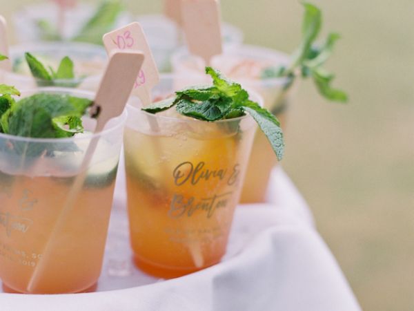 The image shows plastic cups filled with drinks, garnished with mint leaves, and small wooden stirrers, placed on a white cloth.