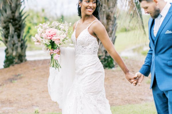 A bride and groom hold hands outdoors, the bride in a white dress holding flowers, groom in a blue suit under palm trees.