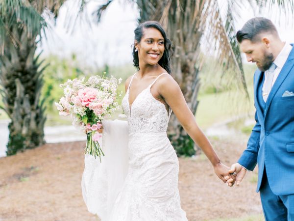 A bride and groom hold hands outdoors, the bride in a white dress holding flowers, groom in a blue suit under palm trees.
