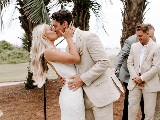 A bride and groom share a kiss outdoors beneath palm trees, with bridesmaids and groomsmen in beige attire standing nearby, during a wedding.