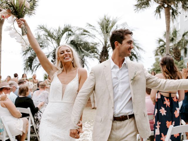 A joyful couple, possibly just married, walks hand in hand among smiling guests, set in a tropical outdoor venue with palm trees.