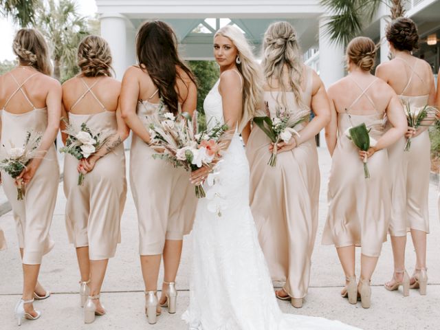 A bride and bridesmaids in light dresses pose for a wedding photo, holding bouquets behind their backs, outdoors.