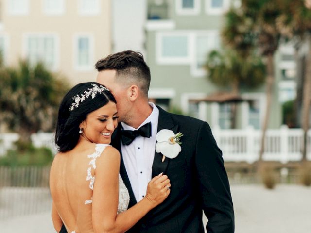 A couple is embracing on a beach, dressed formally, with pastel buildings and palm trees in the background.